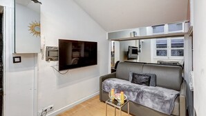 Un espace de vie compact comprenant un canapé, un téléviseur à écran plat fixé au mur, une petite table avec des boissons et une fenêtre équipée de stores.