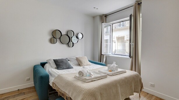 Ein modernes Schlafzimmer mit einem Bett, einem blauen Sofa, einem wandmontierten Spiegel und einem Fenster mit Blick auf Gebäude.