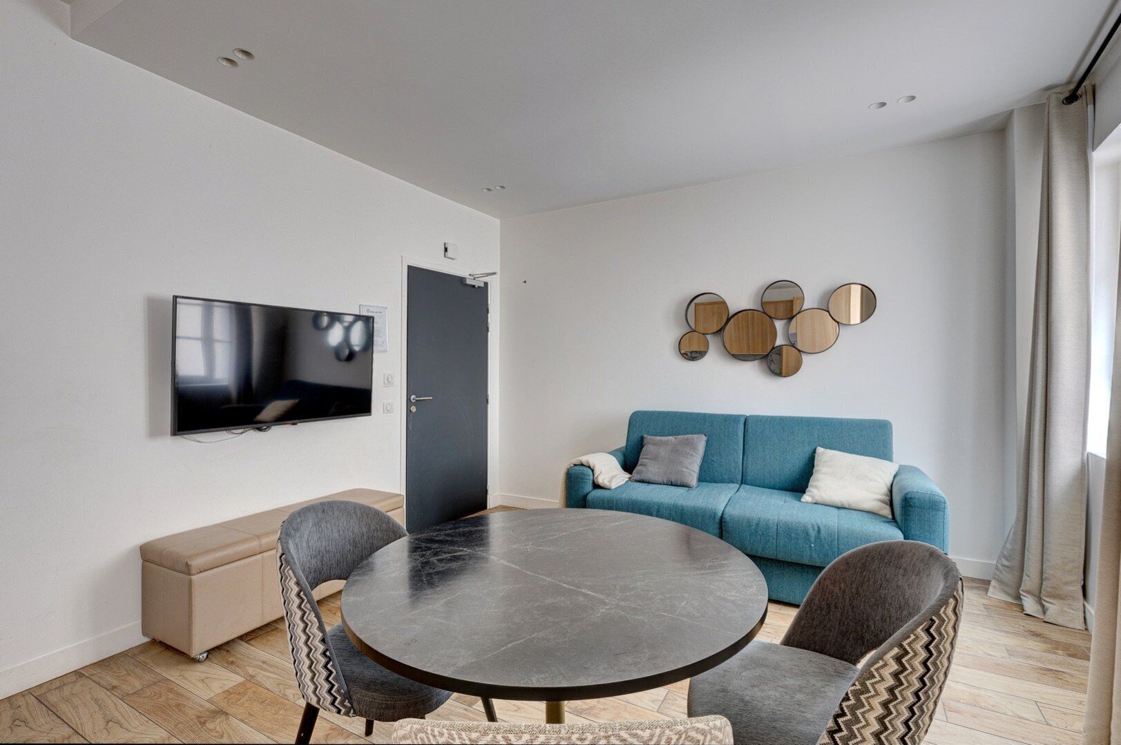 A modern living room with a round table, two armchairs, a blue sofa, a TV, and a wall-mounted mirror.