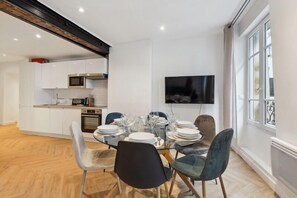 Un espace repas moderne avec une table en verre disposée pour six personnes, entourée de chaises dotées de sièges rembourrés. On aperçoit à l’arrière-plan une cuisine avec des meubles blancs et un four à micro-ondes.