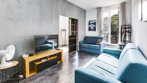 Ein modernes Wohnzimmer mit einer grauen Betonwand, einem blauen Sofa, einem Fernseher auf einem Holzboden und einem großen Fenster mit Vorhängen.