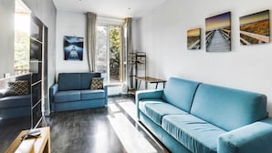 Ein modernes Wohnzimmer mit einem blauen Sofa, einem Couchtisch und einem Fernseher. An der Wand hängen drei Gemälde und ein Fenster mit Vorhängen.