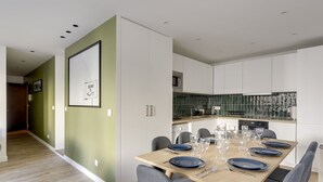 A modern dining area with a wooden table set for six, surrounded by grey chairs. A kitchen with a green backsplash and white cabinets is visible in the background. - 173 Suite Eiffel - Superb apartment in Paris. (Paris 15 Vaugirard)