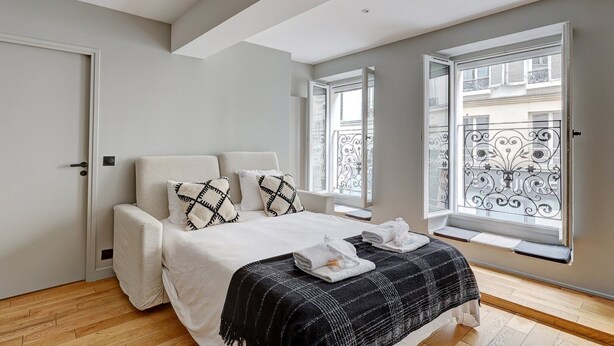 A modern bedroom with a bed, two pillows, a bedside table, and a window with a balcony view. - 169 Suite W - Elegant Apartment in Paris (Paris 02 Bourse)