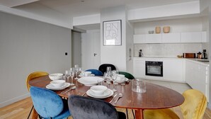 A modern dining area with a wooden table set for six, surrounded by colorful chairs. A kitchen with white cabinets and a black oven is visible in the background. - 169 Suite W - Elegant Apartment in Paris (Paris 02 Bourse)