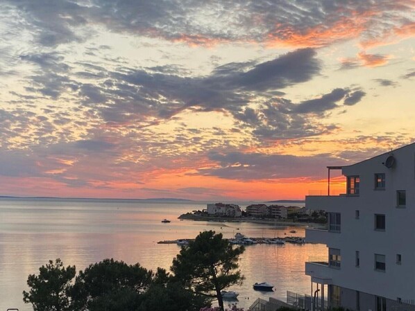 A coastal view with a sunset, calm sea, and a building with balconies overlooking the water.