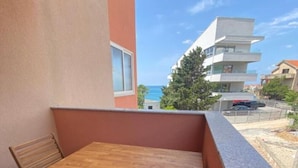 Een balkon met een houten tafel en stoelen, een gewatteerd bankje en uitzicht op zee en gebouwen.