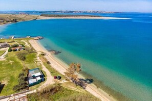 An aerial view of a coastal property with a beach, a fenced area with parked cars, and a building with a green roof.