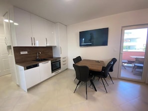 Una cocina moderna con armarios blancos, encimera de madera y horno empotrado. Hay una mesa de comedor con sillas negras y un televisor montado en la pared.