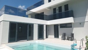 A modern two-story house with a swimming pool, glass doors, and outdoor furniture. - Queen Room with Pool View (Oistins)