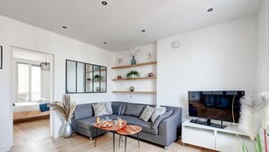 En moderne stue med en grå sofa, et træsofabord og et fladskærms-TV på et hvidt stativ. Reoler med planter og pynteting, samt et vindue med udsigt udad.