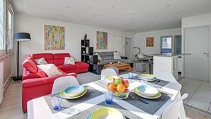 Ein modernes Wohnzimmer mit einem roten Sofa, einem Esstisch mit Geschirr und einer Obstschale, und einer Küchenzeile im Hintergrund.