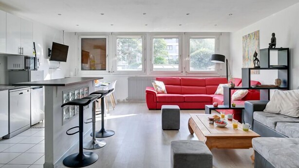 Eine moderne Küche mit weißen Schränken, einer zentralen Kochinsel mit Barhocker, einem roten Sofa und einem Couchtisch mit Frühstücksgebäck.