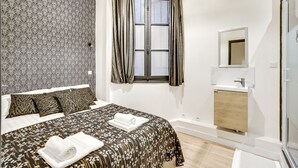 Un dormitorio con cama, ventana con cortinas, espejo en la mesita de noche y baño con lavamanos y espejo.