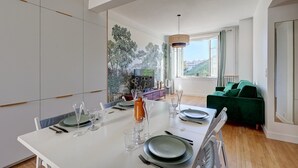 A modern dining area with a white table set for four, a green sofa, and a view of a cityscape through large windows. - 628 Suite Foret - Superb apartment in Paris (Paris 14 Observatoire)