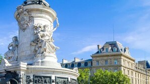 Une statue posée sur un socle circulaire, entourée d’un bâtiment à l’architecture classique.