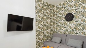 Un salon avec un canapé gris, une petite table ronde sur laquelle se trouvent des fruits et des boissons, un téléviseur fixé au mur et une horloge.