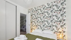 Une chambre d’hôtel avec un lit, une couvre-lit verte, un papier peint aux motifs tropicaux, une table de chevet avec une lampe et une porte donnant sur une autre pièce.