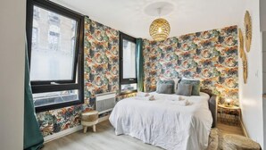 Un dormitori amb un llit gran, paper pintat amb motius florals, una finestra i un llum penjant.