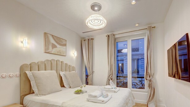 A hotel room with a large bed, a bedside table, a window with curtains, and a chandelier. - 348 Ponthieu - Superb apartment in Paris (Paris 08 Élysée)