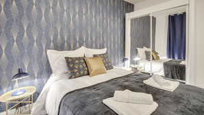 A hotel room with a geometric patterned wallpaper, a large bed with white and gold bedding, a bedside table with a lamp, and a mirror reflecting the room.