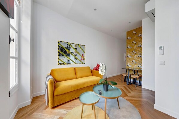 A living room with a yellow sofa, a glass coffee table, and a painting on the wall. - 210 Suite Chanel - Superb apartment in Paris (Paris 04 Hôtel-de-Ville)