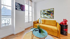 A living room with a yellow sofa, a glass coffee table, and a potted plant. There are two windows with blinds, a painting on the wall, and a red sculpture on a black container. - 210 Suite Chanel - Superb apartment in Paris (Paris 04 Hôtel-de-Ville)