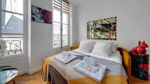 A hotel room with a bed, a glass table, a window with blinds, and artwork on the wall. - 210 Suite Chanel - Superb apartment in Paris (Paris 04 Hôtel-de-Ville)