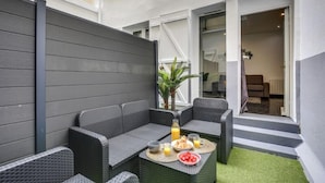 Un espace extérieur moderne aménagé avec un canapé et des fauteuils, une table où sont disposés des éléments pour le petit-déjeuner, et un escalier menant à un espace intérieur.