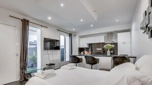 Une chambre d’hôtel moderne dotée d’un grand lit, d’une porte vitrée donnant sur un balcon, d’une kitchenette et d’un coin salon avec deux fauteuils.