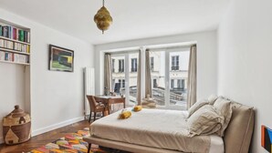 A bedroom with a bed, a desk, a chair, a bookshelf, and a view of buildings through large windows. - 620 Suite Martel - Superb apartment in Paris. (Paris 10 Entrepôt)