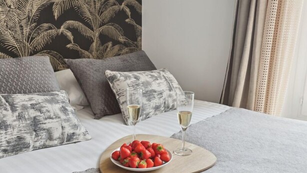 A neatly made bed with a white and gray patterned bedspread, a wooden tray with strawberries, two glasses of champagne, and a tropical leaf-patterned headboard. - 867-Suite Mimosa - Superb Apartment (Montreuil)