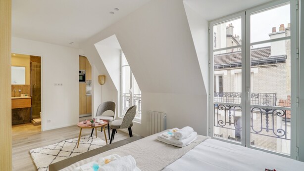 Et moderne soveværelse med et stort vindue, en balkon med gelændere, en seng med hvide sengetøj, et lille rundt bord med en stol og udsigt over bybilledet.