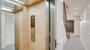 En moderne elevator med digitalt display og kontrolpanel, en trævæg og en trappe med gelænder af træ.