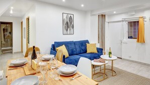 Ein modernes Wohnzimmer mit einem blauen Sofa, einem Esstisch für vier Personen und einem kleinen runden Tisch mit einer Champagnerflasche.