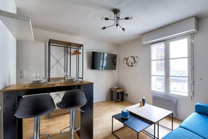 Ein kompakter Wohnraum mit Essbereich, einem an der Wand befestigten Fernseher, einem Fenster mit Blick auf Gebäude und einem blauen Sofa.