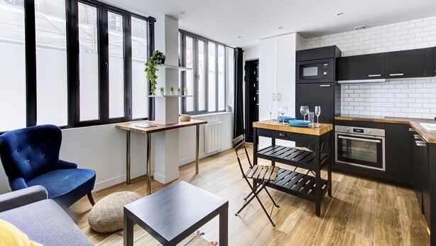 A modern kitchen with black cabinets, a microwave, and an oven. There is a dining table with chairs and a blue armchair. - 318 Suite Pivert - Superb apartment in Paris (Paris 11 Popincourt)