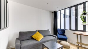 A modern living room with a grey sofa, a blue armchair, a small table, and a window with black curtains. - 318 Suite Pivert - Superb apartment in Paris (Paris 11 Popincourt)