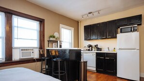 Une cuisine compacte avec des meubles noirs, un réfrigérateur blanc et un four à micro-ondes. On y trouve une fenêtre avec des stores, un petit coin bar avec des tabourets et une poubelle.