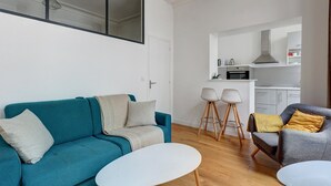 A modern living room with a teal sofa, a white coffee table, and a grey armchair. A kitchen is visible in the background with white cabinets and a stove. - 338 Suite Montmartre - Superb apartment in Paris (Paris 09 Opéra)