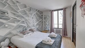 Ein modernes Schlafzimmer mit geometrischem Wanddesign, einem großen Bett mit weißer Bettwäsche und einem Fenster mit gestreiften Vorhängen.