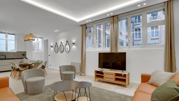 Ein modernes Wohnzimmer mit einer Couch, zwei Sesseln, einem Couchtisch, einem Fernseher und einem Essbereich mit Stühlen und einem Tisch.