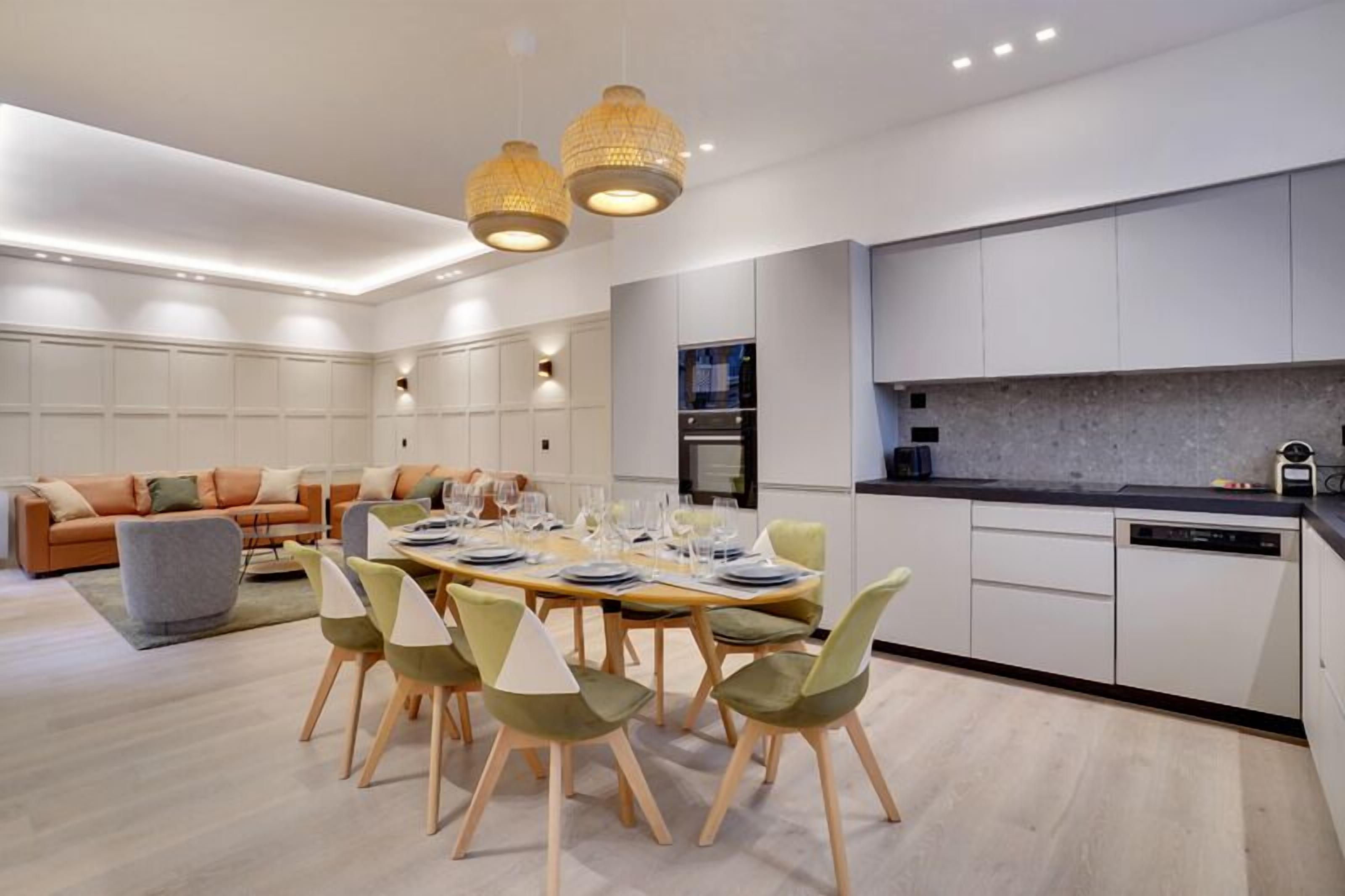 A modern kitchen with white cabinetry, a dining area with a wooden table and chairs, and a living room with a sofa and cushions.
