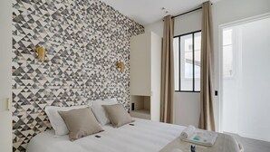 Ein Hotelzimmer mit Wand im geometrischen Muster, einem Bett mit weißen Leinen und einem Fenster mit Vorhängen.