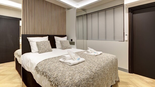 A modern hotel room with a large bed, a wooden slat headboard, a large window with blinds, and a dark door. - 327 Suite Le Marais - Superb apartment in Paris (Paris 03 Temple)