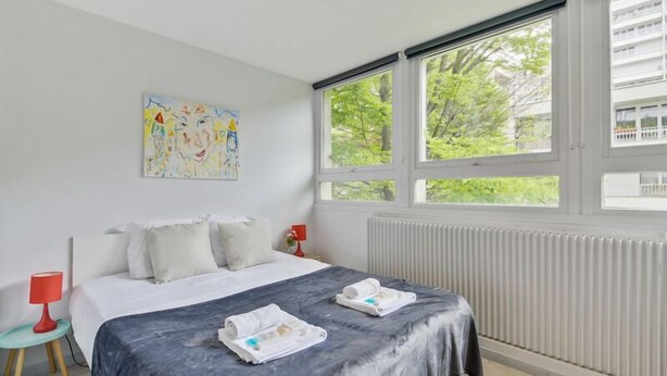 A modern hotel room with a large bed, a window with a view of trees, a radiator, and a colorful abstract painting on the wall. - 233 Suite Home Paris Concept (Paris 20 Ménilmontant)