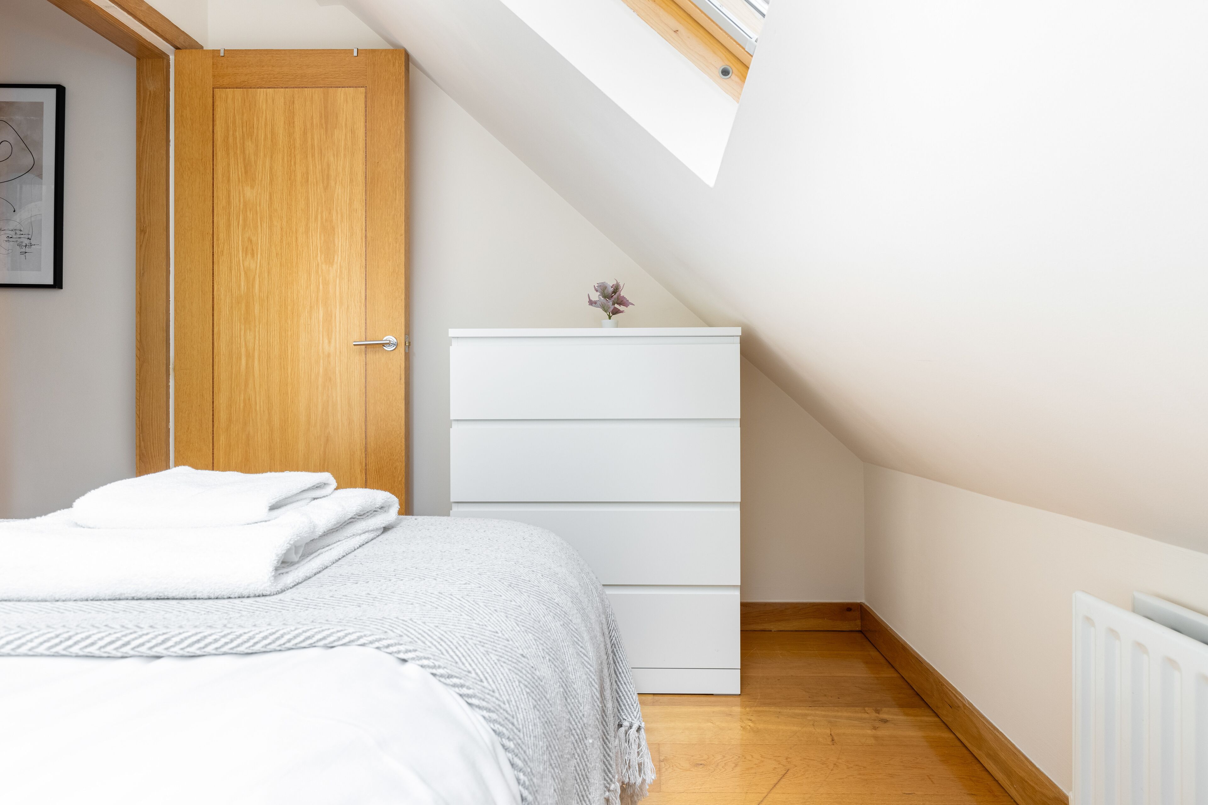 A bedroom with a bed, a white dresser, a wooden door, and a skylight.