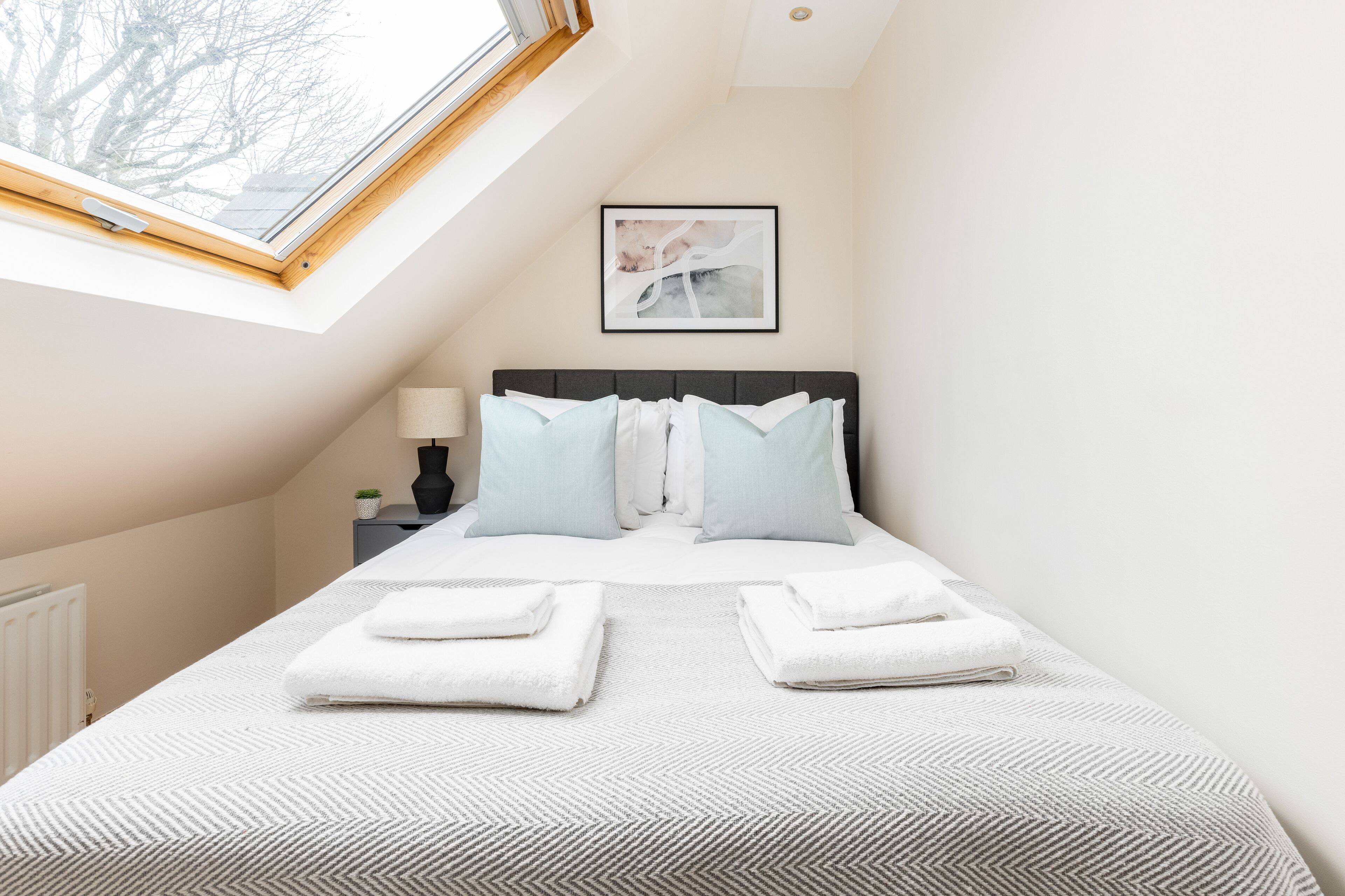 A neatly made bed with pillows and folded towels, a bedside lamp, and a framed abstract artwork on the wall.