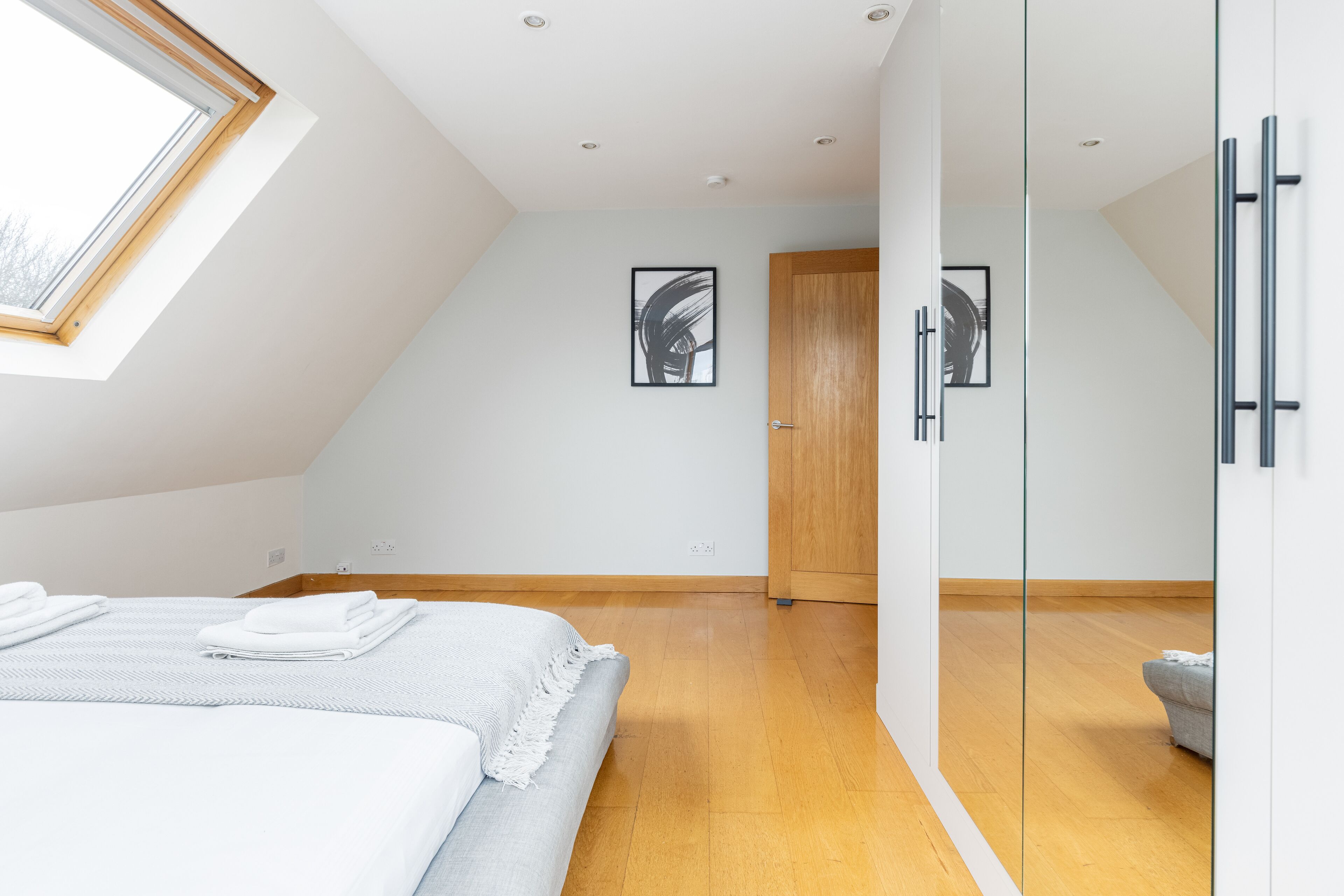 A modern bedroom with a skylight, a bed with neatly folded white towels, a grey sofa, and two framed abstract paintings on the wall.