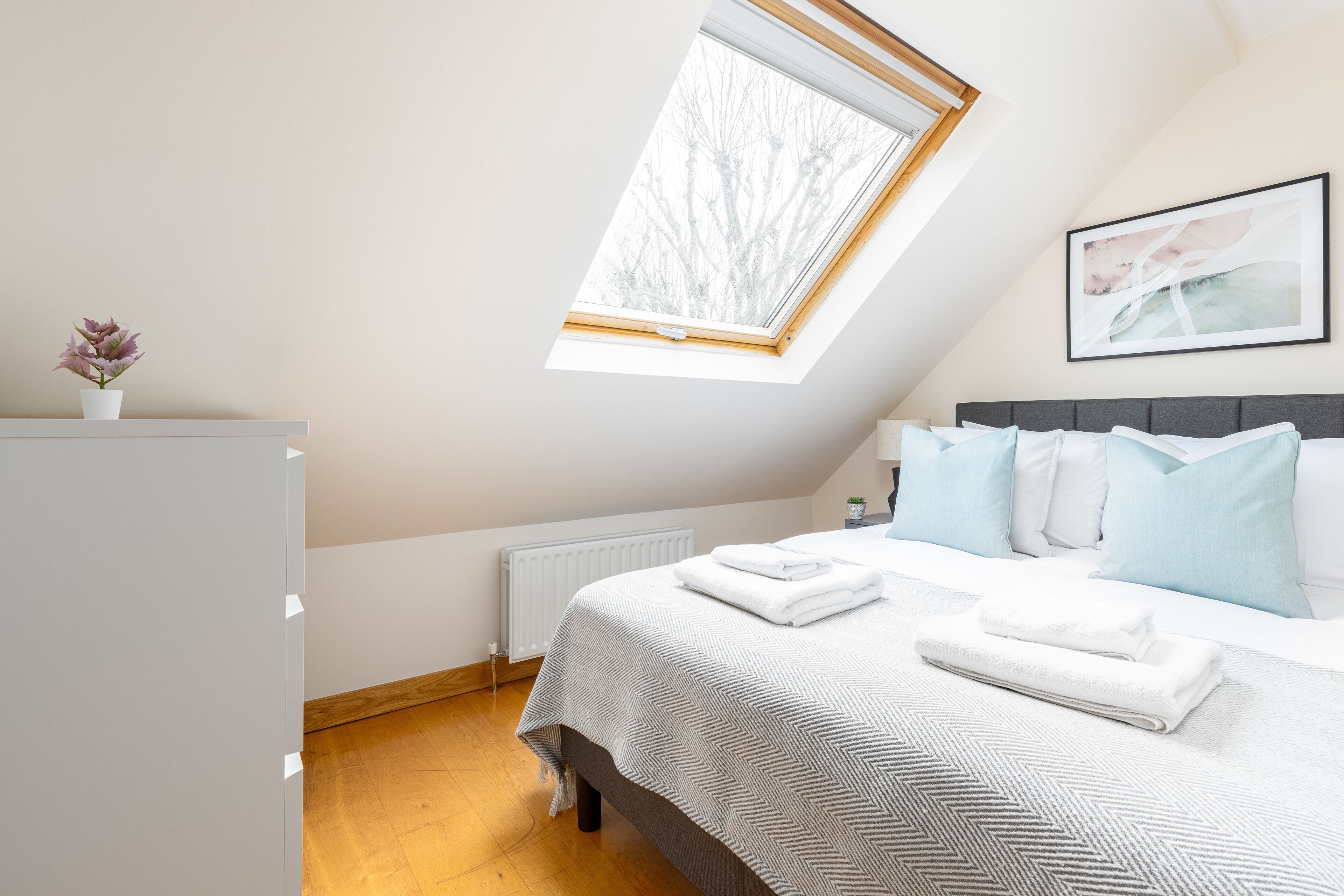 Ein Schlafzimmer mit Oberlicht, einem Bett mit weißer und blauer Bettwäsche, einem Heizkörper und einem gerahmten Bild an der Wand.